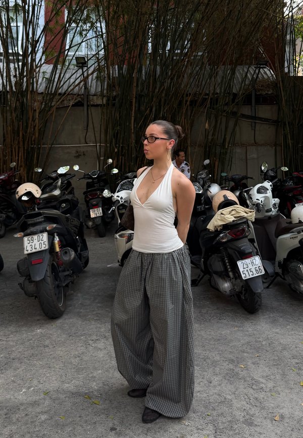 Femme avec des lunettes se tient les mains derrière le dos dans un parking rempli de scooters avec de grands arbres fins en arrière-plan.
