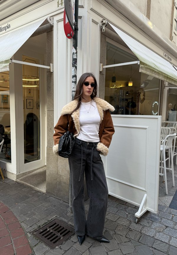 Woman wearing brown shearling jacket, white shirt, black wide-leg jeans, and sunglasses stands on a city pavement near café entrance.