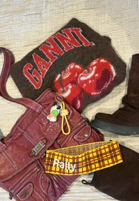 Sac fourre-tout en feutre marron avec des cerises rouges et "GANNI", bottes en suède marron, sac en cuir rouge avec porte-clés, et serre-tête à carreaux jaunes "Rally" sur une surface en tissu.