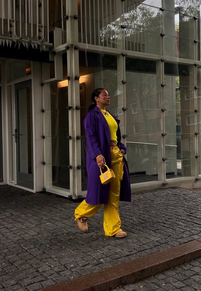 Abrigo largo morado, top y pantalones amarillos, bolso amarillo. Texturas suaves, colores vivos, diseño casual, con un fondo arquitectónico moderno.