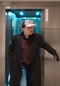 Een persoon in een zwarte jas en een bruin gebreide trui met patroon staat in een zilverkleurige lift, draagt een spijkerpet en zwarte handschoen, met blauw licht.