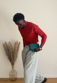 Borsa a tracolla rettangolare color turchese con chiusura a zip, indossata in vita. Modello con dolcevita rossa e pantaloni grigio chiaro, accanto a una pianta alta.