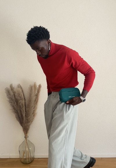 Bolso cruzado rectangular en color teal con cierre de cremallera, llevado en la cintura. Modelo con un suéter de cuello alto rojo y pantalones gris claro, junto a una planta alta.