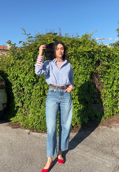Mujer con cabello largo y oscuro está junto a una pared densa de enredaderas verdes, vistiendo jeans azules, una camisa azul abotonada sobre una camiseta de rayas rojas y zapatos planos rojos.
