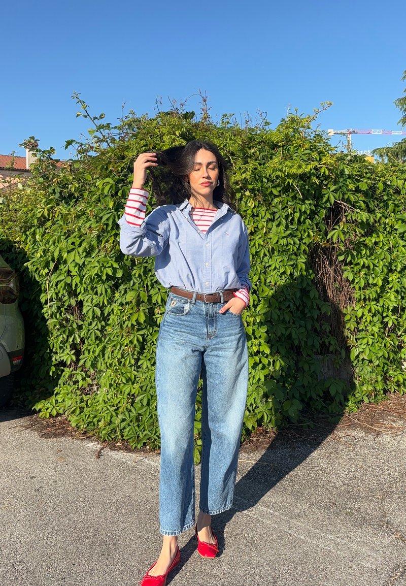 Femme aux longs cheveux foncés debout près d’un mur dense de vigne verte, portant un jean bleu, une chemise bleue boutonnée par-dessus un haut rayé rouge, et des ballerines rouges.