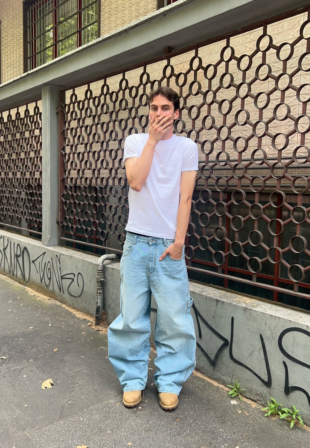 Jeans de ganga largos azuis, t-shirt branca de manga curta e botas de trabalho castanhas. Em pé ao lado de uma cerca metálica decorativa com uma parede coberta de graffiti.