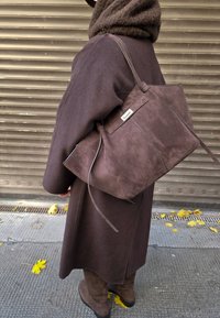 Barna színű, téglalap alakú szarvasbőr válltáska oldalsó pántokkal és fém díszítéssel. Sötét kabáton hordva, texturált háttér előtt.