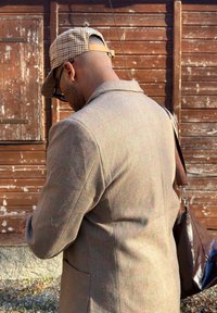 Homme portant une casquette à carreaux, des lunettes et une veste marron à carreaux, portant un sac à bandoulière marron foncé, debout devant un mur en bois.