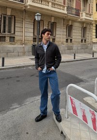 Veste noire zippée portée sur une chemise bleu clair, associée à un jean en denim bleu et des chaussures noires. En arrière-plan, un bâtiment en brique et des barrières de rue.