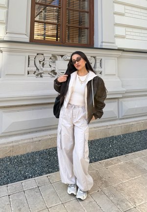 Mujer con cabello largo y oscuro, con gafas de sol, chaqueta marrón, pantalones blancos y zapatillas, de pie sobre un pavimento de baldosas junto a un edificio blanco ornamentado.
