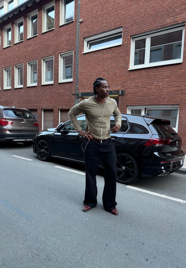 Homme avec des écouteurs debout dans la rue à côté d'une voiture noire garée devant un bâtiment en briques rouges avec plusieurs fenêtres.