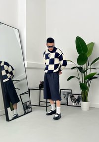 Marineblauw en crèmekleurig geblokt gebreid sweater, geplooide donkere shorts, witte sokken, zwarte schoenen, met een spiegel en een potplant op de achtergrond.