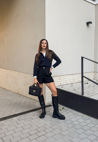 Cárdigan negro de punto trenzado sobre una camisa blanca, combinado con pantalones cortos negros de tiro alto. Botas negras hasta la rodilla y un pequeño bolso negro completan el look.