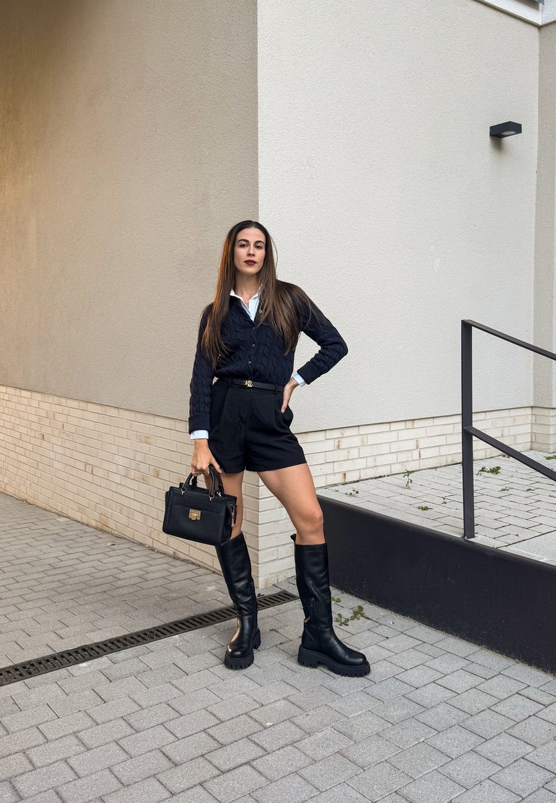 Cardigan de malha canelada preta sobre uma camisa branca, combinado com calções pretos de cintura alta. Botas pretas até ao joelho e uma pequena carteira preta completam o visual.