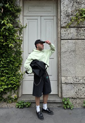 Sudadera oversized verde claro, pantalones cortos negros a rayas, gorra negra y bolsa negra, con calcetines y zapatos blancos. Fondo de follaje verde y pared de piedra.