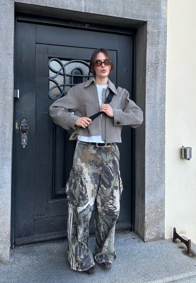 Mujer joven con gafas de sol, chaqueta beige de gran tamaño, pantalones cargo de camuflaje, ajustando la correa de un bolso negro al hombro, de pie frente a una puerta negra.