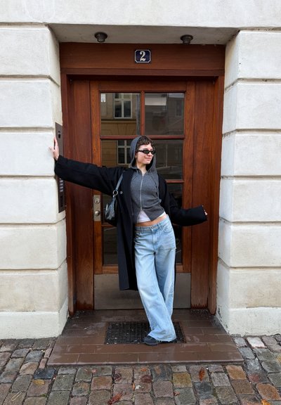 Persona con gafas de sol y chaqueta con capucha apoyada con los brazos extendidos frente a una puerta de madera con el número 2 en un edificio de piedra.