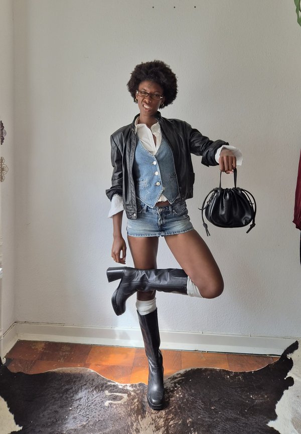 Femme portant une veste en cuir noire, un gilet en denim et un short, des bottes noires montant jusqu'aux genoux, tenant un sac à main noir, debout sur un tapis en peau de vache.