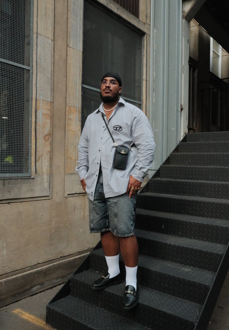 Ριγέ πουκάμισο μακρύ μανίκι, τζιν σορτσάκι, μαύρη τσάντα ώμου, λευκές κάλτσες, μαύρα loafers, στέκεται σε ένα μεταλλικό σκαλοπάτι μπροστά σε έναν υφή τοίχο.