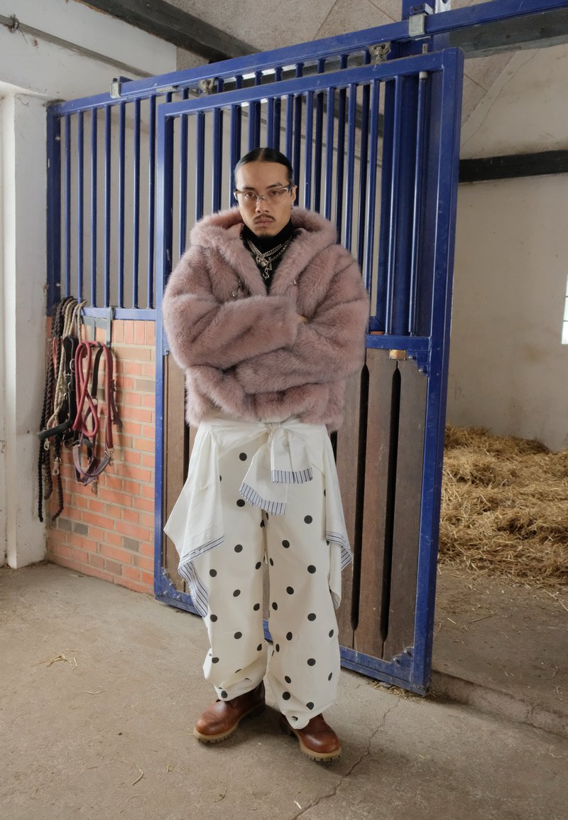 Man i rosa pälsjacka, vita byxor med prickar och bruna stövlar som står med armarna i kors i en stallbyggnad nära en blå metallgrind.