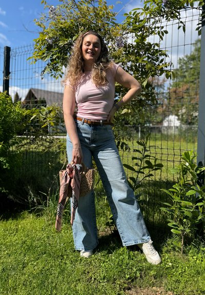 Una mujer de pie al aire libre con una camiseta ajustada rosa y unos jeans de pierna ancha azul claro, sosteniendo una cesta de mimbre con una bufanda estampada. Hay un fondo de follaje verde.
