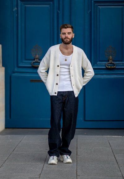 Cárdigan blanco de canalé con botones oscuros sobre una camiseta blanca, combinado con jeans de mezclilla oscuros y zapatillas blancas, frente a una puerta azul.