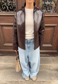 Veste en cuir synthétique brun avec un col haut, fermeture éclair à l'avant, portée par-dessus un haut clair et un jean bleu clair ample. Accentuée par un sac à motifs.