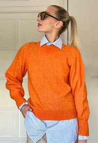 Oranž plekk-sviiter soonikute ja äärtega, kihiliselt sinise ja valge triibulise kaelusega särgi peal. Sujuv tekstuur, mugav lõige.