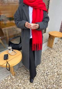 Manteau en laine gris, écharpe rouge à franges, chemise blanche, pantalon noir, tenant une tasse. Sac noir, verre d'eau, table en bois avec un petit appareil photo à proximité.