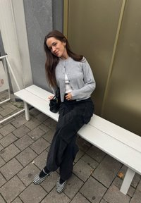Cardigã cinza com nervuras sobre uma blusa branca, calças de carga pretas e sapatos pretos com tachas brancas. Sentada num banco branco contra um fundo neutro.