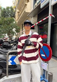 Striped long-sleeve top in maroon and cream, grey jogging bottoms, black gloves, and a cap, with a shoulder bag. The background shows motorcycles and signage.