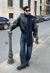 Veste aviateur en cuir noir, écharpe bleue marine, jean décontracté en denim bleu et bottines noires. Debout à côté d'un poteau de rue dans un cadre urbain.