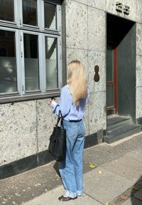 Μαύρη τσάντα, μπλε ριγέ πουκάμισο, ανοιχτό μπλε τζιν παντελόνι, στέκεται κοντά σε ένα λιθόκτιστο κτήριο με μεγάλες βιτρίνες και μια χάλκινη πλάκα.