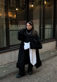 Sort lang frakke med pelskanter, sort top, løse sorte bukser, hvid skjorte bundet foran og sort tekstureret taske. Kraftige sorte sko og sølverkæde.