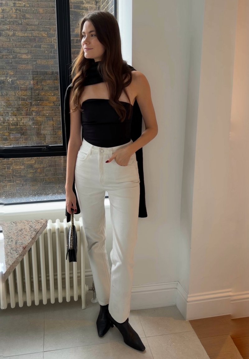 Femme aux longs cheveux bruns debout à l'intérieur, portant un haut noir sans bretelles, un pantalon blanc taille haute, des bottes noires, tenant un sac noir près d'une fenêtre.