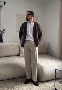 Homme portant un cardigan marron, un t-shirt blanc, un pantalon beige et des chaussures foncées, debout dans un salon aux tons neutres près d'un canapé et d'un tapis beiges.