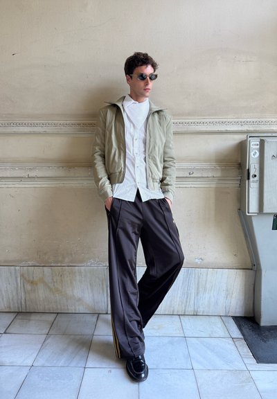 Chaqueta bomber verde claro, camisa blanca a rayas, pantalones anchos oscuros con rayas doradas y zapatos negros. Texturas suaves y diseño moderno.