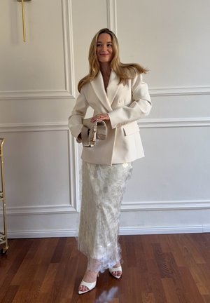 Mujer con blazer blanco y falda larga texturizada sosteniendo un bolso beige, de pie sobre suelo de madera frente a una pared blanca con molduras decorativas.