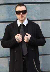 Manteau noir à boutons croisés, chemise blanche à col, cravate noire et bagues aux doigts. Lunettes de soleil noires et détail de chaîne visibles.