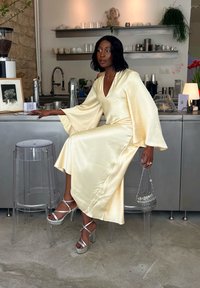 Un vestito in satin di un giallo pallido con maniche a campana, abbinato a scarpe con plateau argentate e lucenti. La modella tiene in mano una borsa metallica con perline.