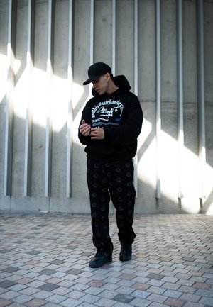 Sudadera negra con diseño gráfico blanco, pantalones negros con un sutil patrón floral y zapatillas negras sobre un suelo de piedra con patrón.