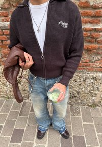 Sötétbarna bordázott cipzáras pulóver fehér logóval, fehér póló, elhalványult kék farmer, barna bőrtáska, ezüstgyűrűk, átlátszó poharat tart a kezében.