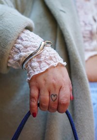Pulseira prateada ondulada sobre uma manga de renda branca; a mão segura uma alça azul, com esmalte vermelho nas unhas e uma tatuagem de coração no dedo.