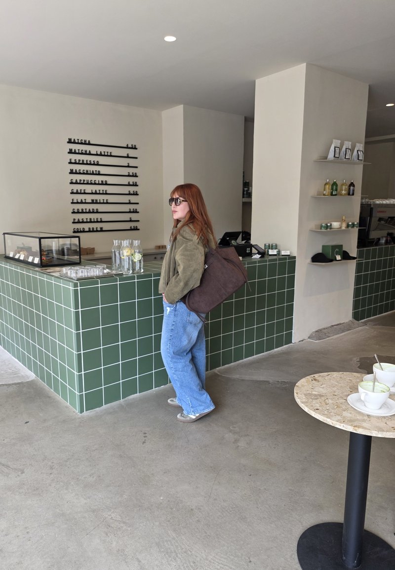 Coffee shop scene featuring a green tiled counter with a glass display, wooden shelves, and a person wearing a brown bag and oversized jeans.