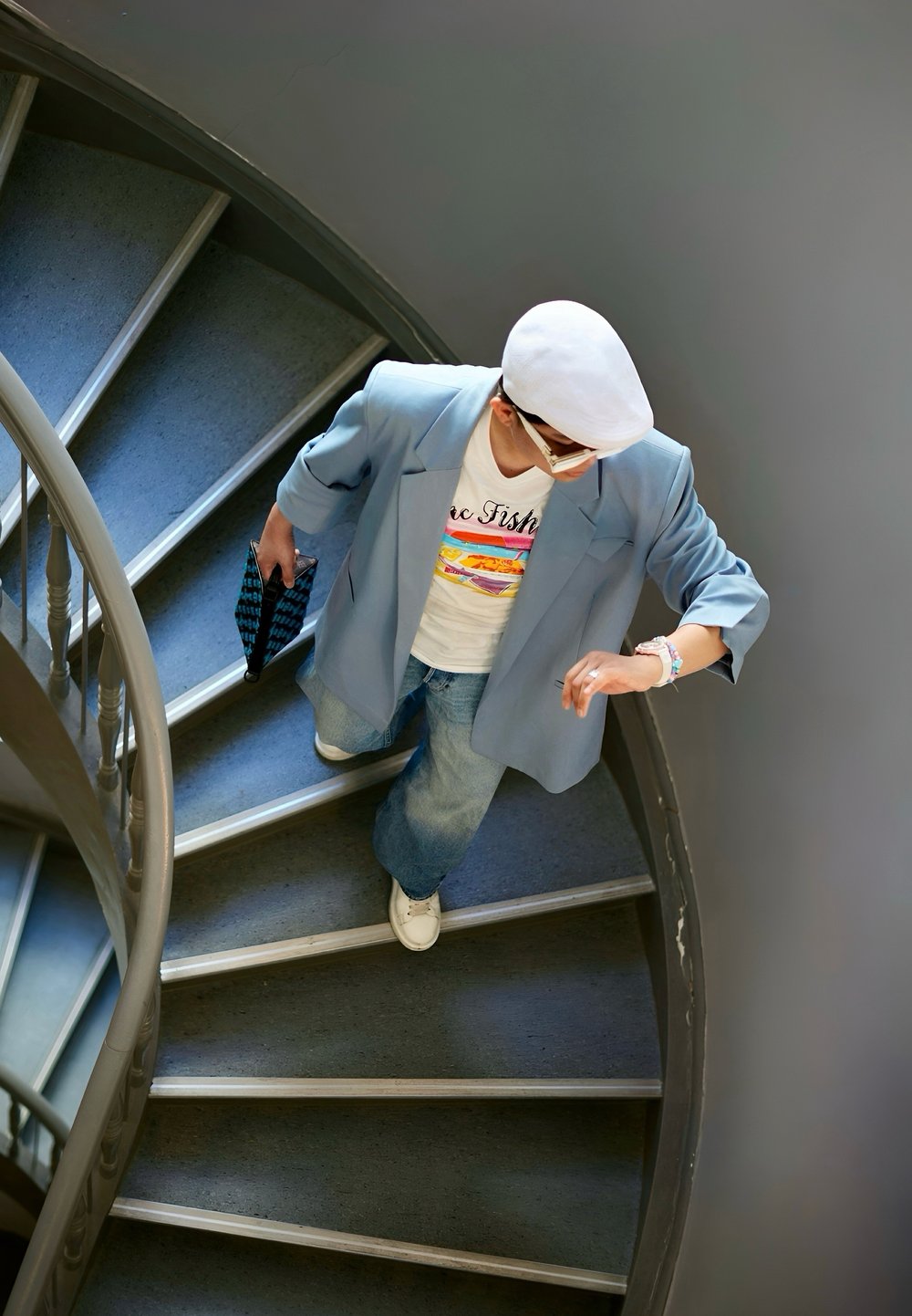 Blazer azzurro, t-shirt bianca con grafica multicolore, jeans di denim larghi, sneakers bianche, che tiene una pochette nera a fantasia, mentre sale le scale.