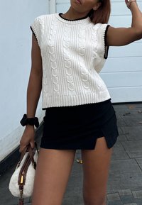 Top senza maniche bianco lavorato a maglia con motivo a treccia e rifiniture nere, abbinato a una mini gonna nera con uno spacco laterale. La borsa è bianca con dettagli marroni.