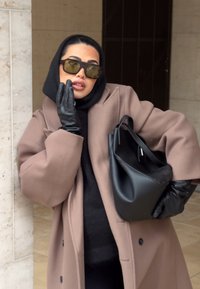 Femme portant un sweat à capuche noir, un pardessus marron, des gants en cuir noirs et des lunettes de soleil, tenant un sac à main en cuir noir, posant près d'un mur en pierre.
