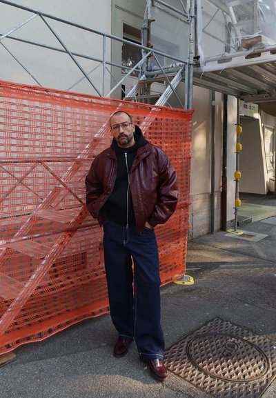 Hombre con chaqueta de cuero marrón, sudadera con capucha negra y vaqueros anchos, de pie con las manos en los bolsillos junto a una valla naranja de construcción en una calle de la ciudad.