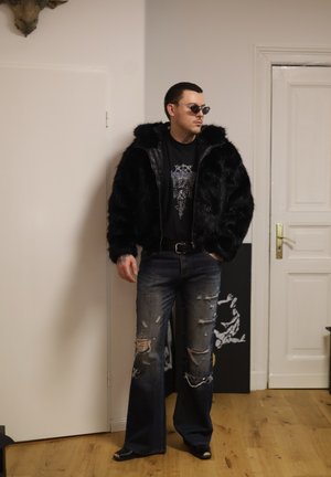 Hombre con chaqueta de piel sintética negra, camiseta gráfica negra, jeans anchos rasgados, gafas de sol, de pie en el interior cerca de una puerta blanca cerrada.