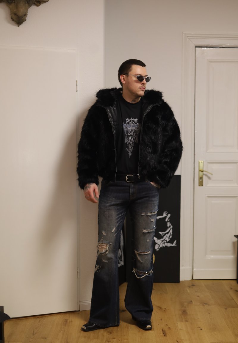 Muž v čiernej bunde z umelého kožušiny, čiernom tričku s potlačou, roztrhaných širokých džínsach, so slnečnými okuliarmi, stojí v interiéri pri zatvorených bielych dverách.